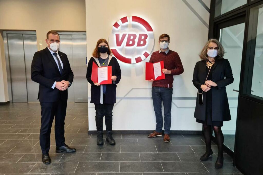 Vier Personen stehen vor einer Wand mit dem VBB-Logo. Die zwei Studierenden halten ihren Vertrag in der Hand