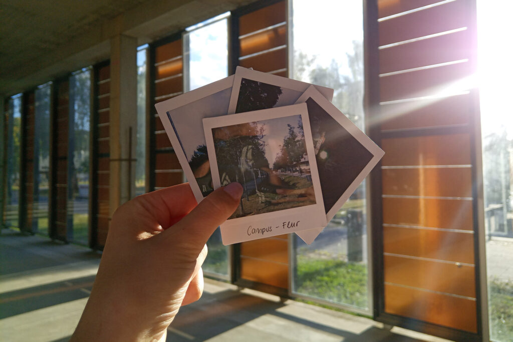 Eine Hand hält vier Polaroids, nur eins ist im Vordergrund auf dem der Campusflur ist. Der Hintergrund ist der sonnendurchflutete Solarpavillion.